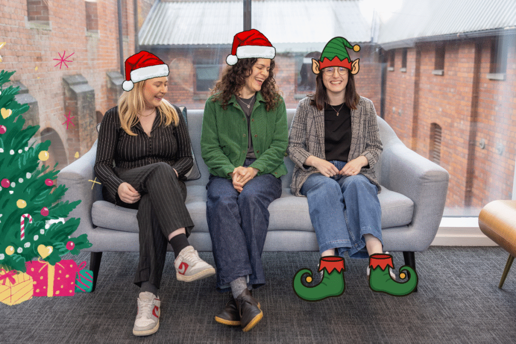 becca, alice and lois sitting on a sofa smiling and laughing at lois as her feet dont touch the floor. with cartoon xmas hats on becca and alice. lois with cartoon elf hats and elf shoes